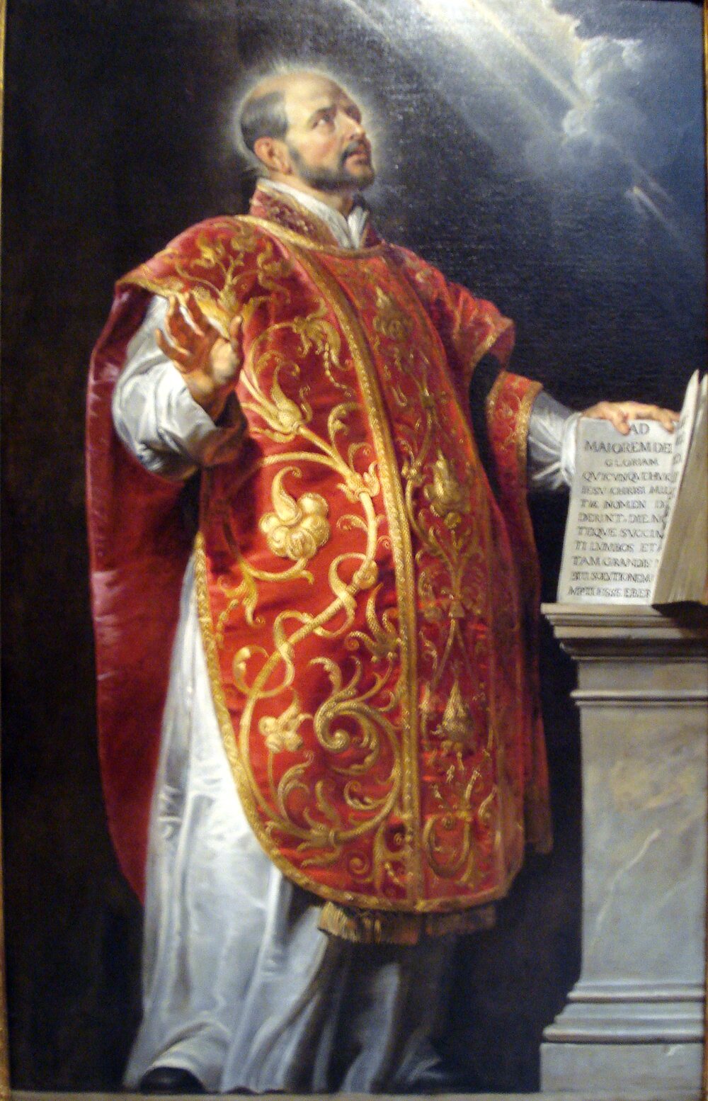 Saint Ignatius of Loyola in a red and gold chasuble, looking up into the light, his hand resting on an open book inscribed Ad Maiorem Dei Gloriam