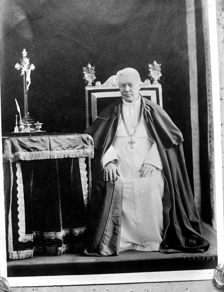Photograph of Pope Saint Pius X seated on the throne in a white cassock beside a prie-dieu